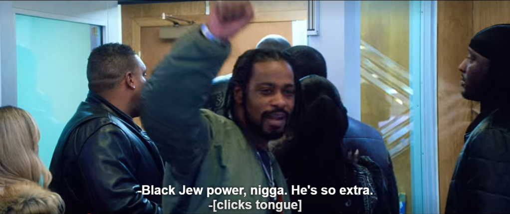 Demany faces the camera as his entourage leaves through the security door. His fist is raised. Subtitle: "-Black Jew power, nigga. He's so extra. -[clicks tongue]"