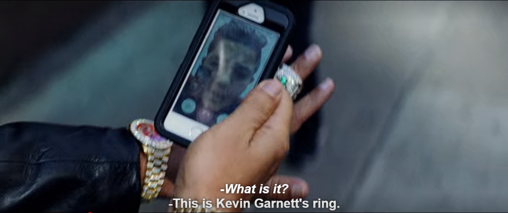 A close-up of Ratner's phone and both his hands. The phone is centered and on it is visible a boy, but light reflections/glare render it difficult to see detail. Ratner holds the phone in his right hand and the phone is framed by a gold watch and the green Celtics ring. Subtitle: (italicized) "-What is it?" (non-italicized) "-This is Kevin Garnett's ring."
